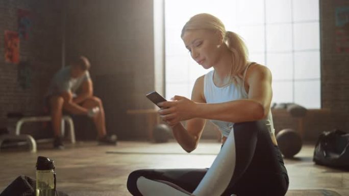 快乐和微笑美丽的运动年轻女子坐在阁楼健身房的地板上时使用智能手机。她在输入信息并思考。一个人在后台锻