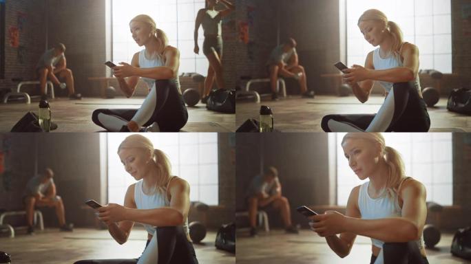 快乐和微笑美丽的运动年轻女子坐在阁楼健身房的地板上时使用智能手机。她在输入信息并思考。一个人在后台锻
