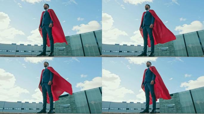 拥有红色斗篷的商人超级英雄站在摩天大楼的屋顶上，准备进行商业交易并节省一天的时间。低角度拍摄。