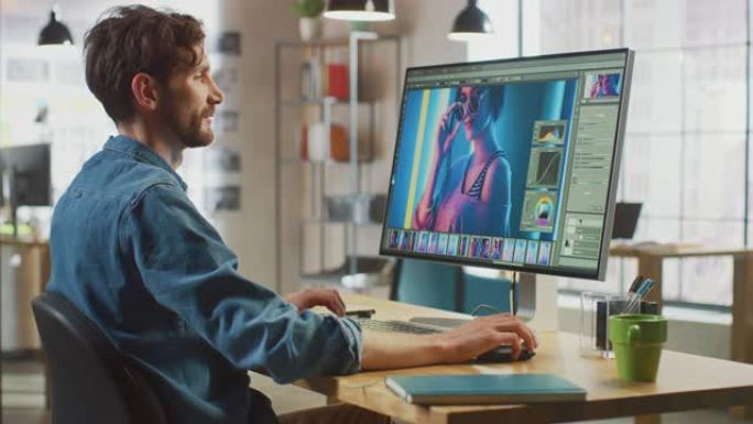 穿着牛仔裤衬衫的男性数字编辑器在他的个人电脑上大显示屏的图像编辑软件中工作。他在凉爽的办公室阁楼工作