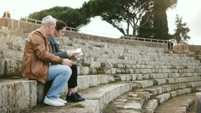 快乐的欧洲高级男人和年轻女子坐在意大利奥斯蒂亚的旧圆形剧场废墟上，带着地图和智能手机。