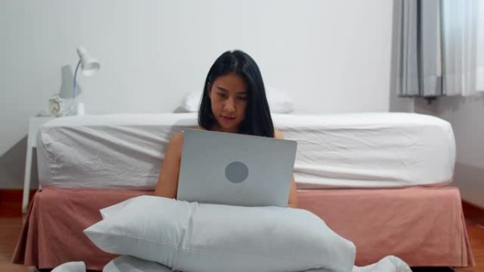 年轻的亚洲妇女使用笔记本电脑检查社交媒体感到高兴的微笑，躺在床上后早上在家里醒来，迷人的泰国女孩微笑