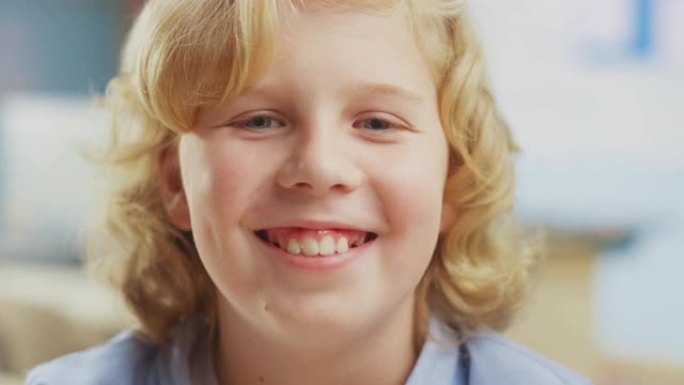 一个可爱的小男孩的肖像，一头金色的卷发坐在他的课桌前，开心地微笑着。聪明的小男孩，带着迷人的微笑坐在
