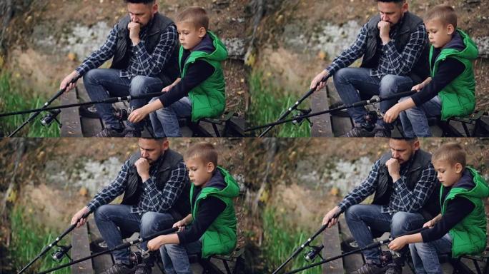 严肃的小男孩正在抓鱼，他的父亲留着胡子的黑发坐在一起，拿着钓鱼竿。共同的爱好、世代和家庭观念。