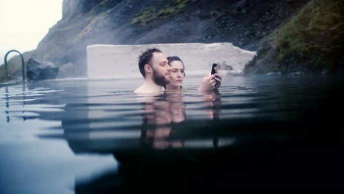 年轻美丽的夫妇在冰岛的温泉游泳。旅行男女在智能手机上拍照。