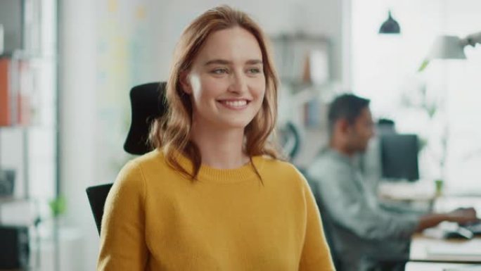 穿着黄色毛衣的红头发的美丽年轻女子的肖像与同事聊天，对着镜头微笑着转身。在明亮多样的办公室工作的成功