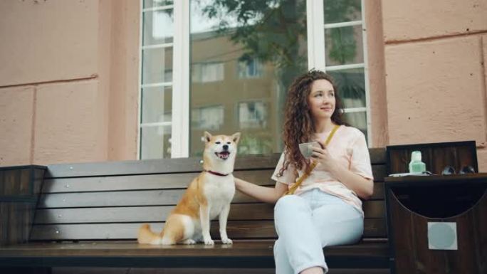 年轻的女士抚摸着坐在咖啡馆外面长凳上的狗，手里拿着一杯咖啡