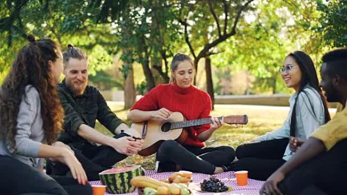 当美丽的年轻女子在公园野餐时弹吉他时，欢快的人在唱歌和大笑的慢动作。音乐、休闲和有趣的概念。