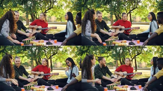 当美丽的年轻女子在公园野餐时弹吉他时，欢快的人在唱歌和大笑的慢动作。音乐、休闲和有趣的概念。