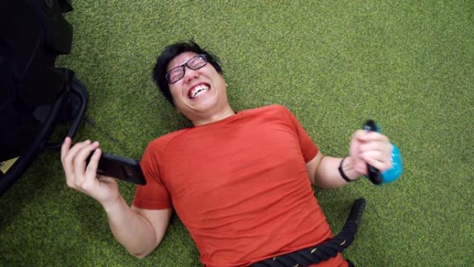 懒惰的亚洲男子躺在背上，在健身房用智能手机看电影
