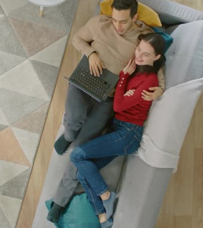 年轻美丽的夫妇躺在沙发上，用笔记本电脑。他们看着上面微笑。顶视图。垂直屏幕方向9:16的视频素材