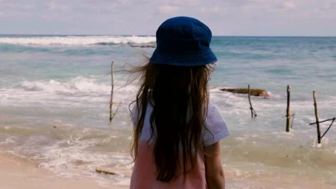 后视图快乐的6-8岁长头发戴帽子的小女孩在异国情调的海洋海滩观看美丽的放松风景