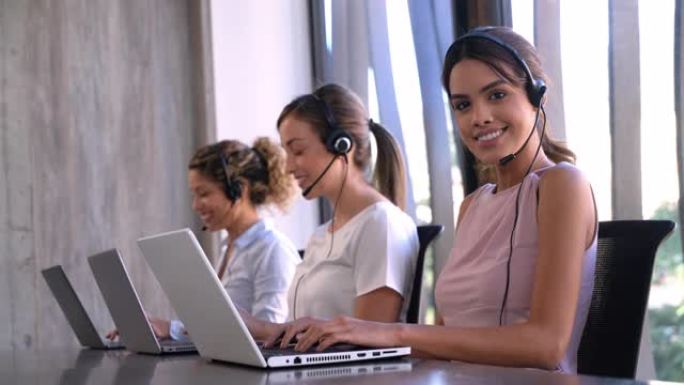 在呼叫中心工作的高效女性在笔记本电脑上打字信息，其中一位看着相机微笑