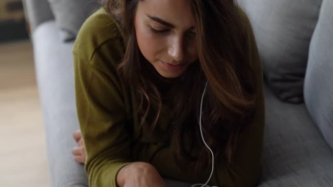 女人躺在肚子上的沙发上，一边看平板电脑上的照片，一边用耳机听音乐