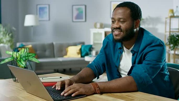 英俊微笑的黑人男子坐在舒适的客厅的桌子旁，用笔记本电脑。