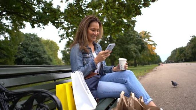 女人从疯狂购物中休息，坐在公园的长椅上，用智能手机发短信，喝 “外卖” 咖啡
