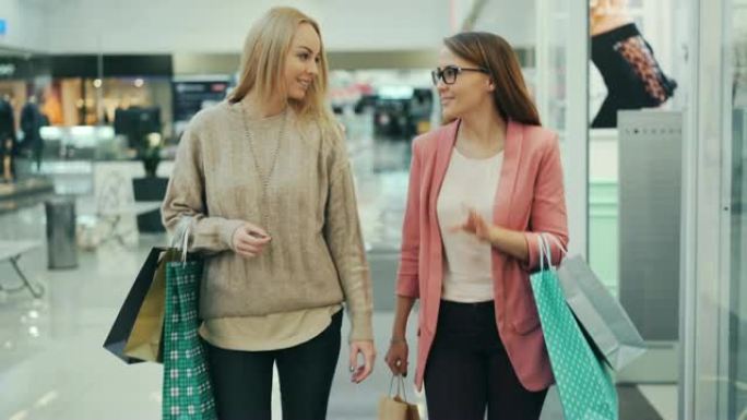 开朗的女人在商店购物中心里提着纸袋一起散步时说话和打手势。女孩们穿着时髦的服装和配饰。