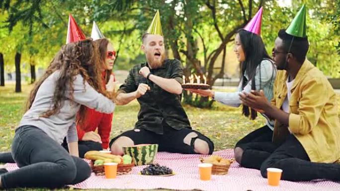 大胡子的男人正在公园里与朋友一起庆祝生日，在蛋糕上吹着蜡烛，喊着并表达积极的情绪，而朋友则在笑着拍手