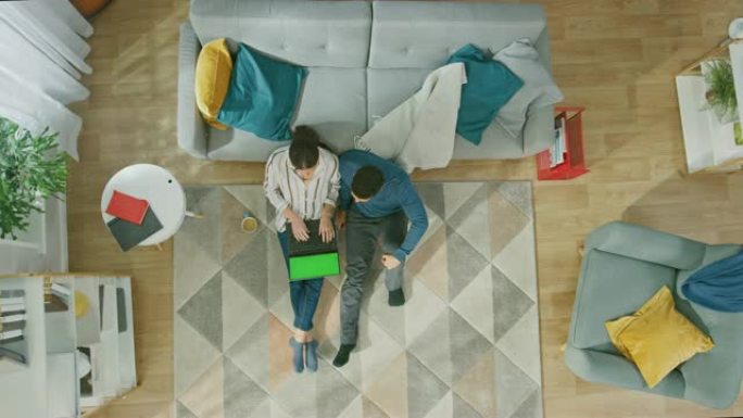 年轻夫妇坐在地板上，使用带有绿屏的笔记本电脑。女孩喝咖啡。舒适的客厅，配有现代室内地毯、沙发、椅子、