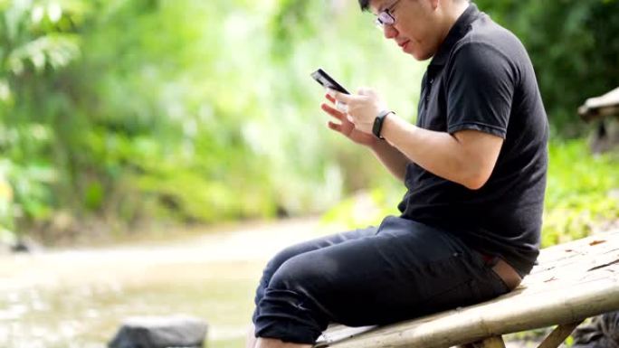 向上倾斜 -- 亚洲男子在典型的河流放松玩智能手机