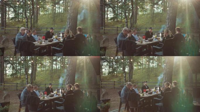 一群朋友坐在松树林中避暑别墅露台上的一张大桌子旁。浪漫的夏日氛围。男孩和女孩喝葡萄酒和其他轻度酒精饮