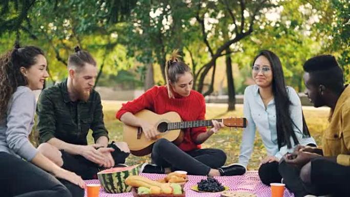 漂亮的女吉他手在野餐时弹吉他，她的朋友们的学生们坐在公园的毯子上唱歌和拍手。露天派对和音乐概念。