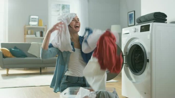 美丽快乐的黑发年轻女子穿着家常牛仔裤的衣服坐在洗衣机旁边。她笑着把洗衣扔到空中。明亮宽敞的客厅，室内