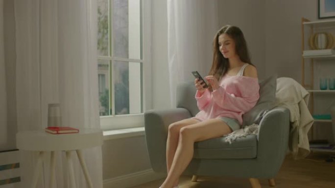 美丽的年轻女子坐在椅子上微笑着使用智能手机。性感的女孩穿着可爱的粉色毛衣和短裤，上网冲浪，在社交媒体