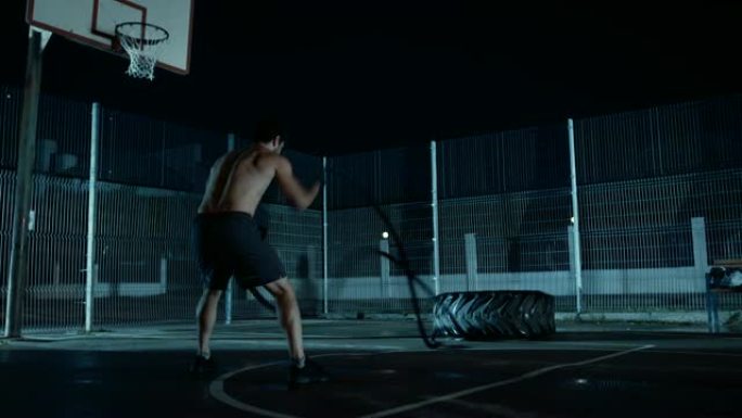 一个强壮的肌肉健康的光着膀子的年轻人用战斗绳做练习的背景。他正在一个有围栏的室外篮球场里锻炼身体。居