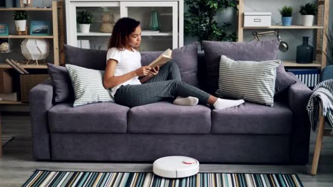 放松的非洲裔美国女孩正在沙发上看书，而机器人真空吸尘器则在家里而不是人的地板上徘徊。小工具和家庭概念