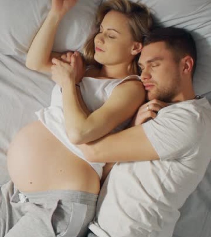 幸福的年轻夫妇与孕妇一起睡在床上，甜蜜的年轻人在睡觉时互相抱着。垂直屏幕方向9:16的视频素材