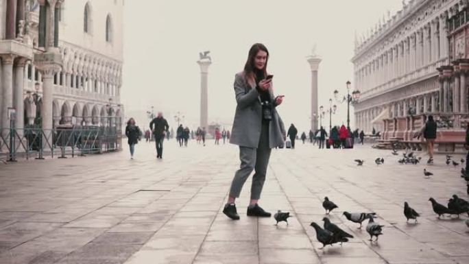 快乐微笑的旅游女孩拍摄智能手机照片站在意大利威尼斯城市广场上的鸽子群慢动作