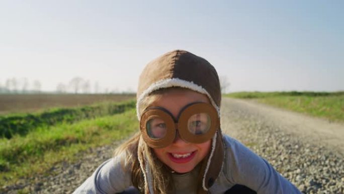 可爱的小女孩的真实照片，戴着飞行员帽子，在春天的自然背景下在镜头前玩得开心，微笑
