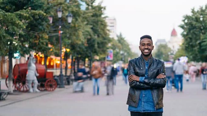 微笑的非洲裔美国男子独自站在市中心，双手交叉，享受城市生活，看着相机。青年和生活方式概念。