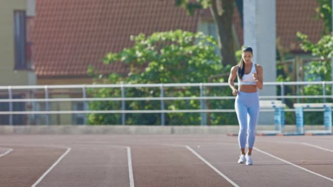 穿着浅蓝色运动上衣和打底裤的美丽健身女子在体育场慢跑。她在一个温暖的夏日下午跑步。运动员在赛道上进行