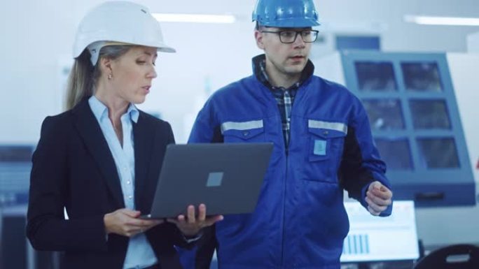 女经理和项目工程师戴着安全帽穿过工业工厂，使用笔记本电脑，交谈，计划生产率优化。生产线工人操作数控机