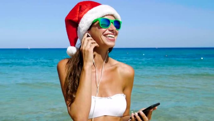 美丽的女孩在手机上自拍，戴着圣诞老人的帽子，戴着墨镜，穿着白色泳衣和白色耳机，背景为海蓝色的水。