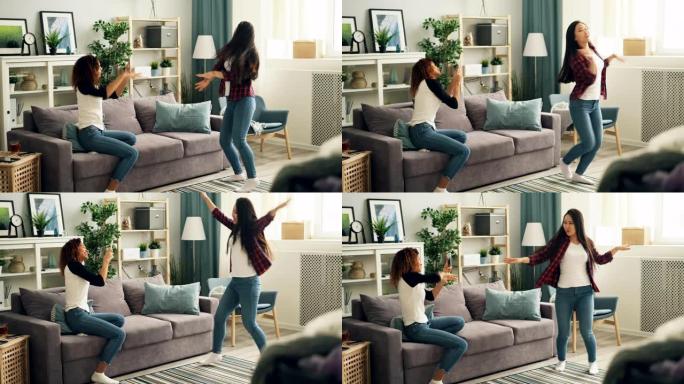 美丽的亚洲女孩穿着休闲服装时尚衬衫和牛仔裤在家里跳舞，而她的非裔美国朋友正在使用智能手机制作视频。