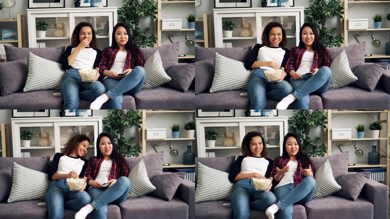 快乐的女性朋友正在一起看有趣的电影，坐在家里的沙发上大笑，交谈和吃爆米花。友谊和电视概念。