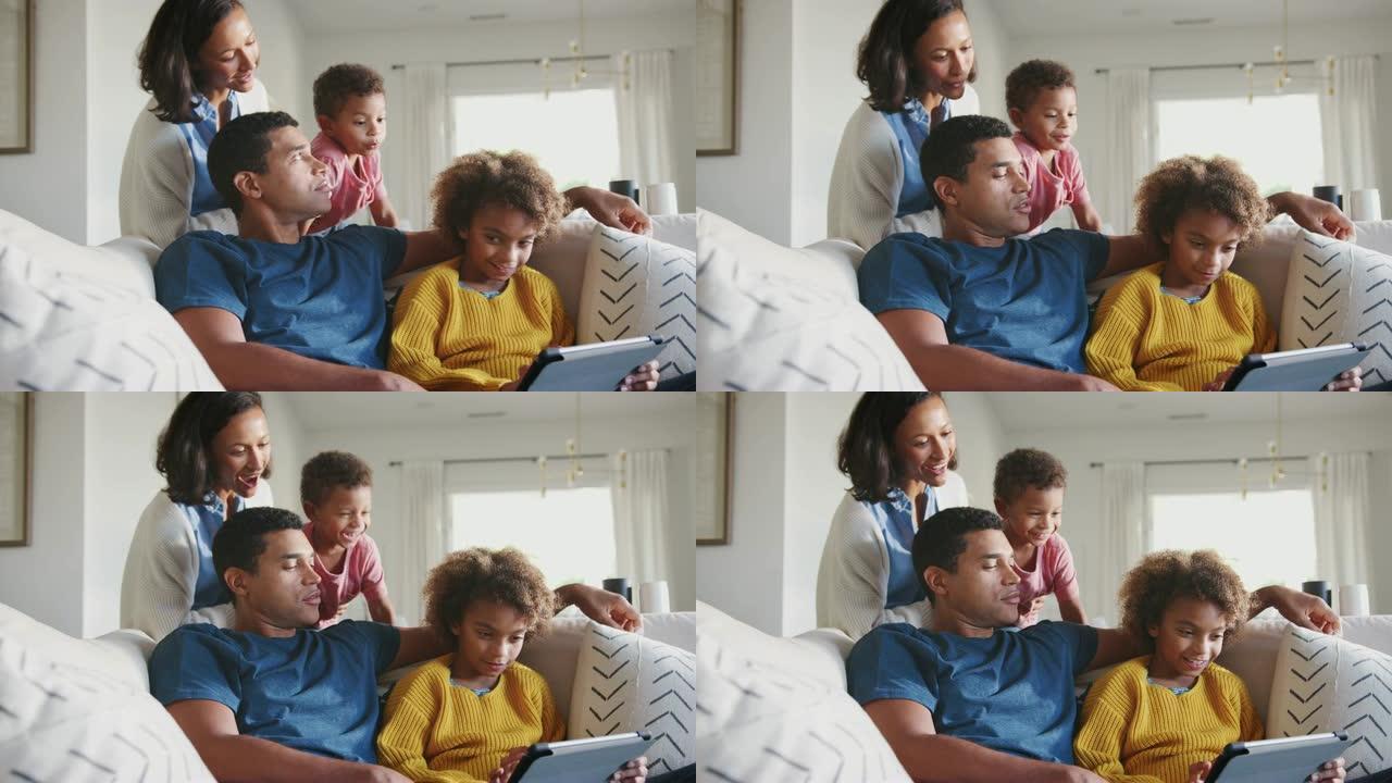 爸爸和女儿在沙发上使用平板电脑，妈妈站在他们身后抱着小孩，低角度