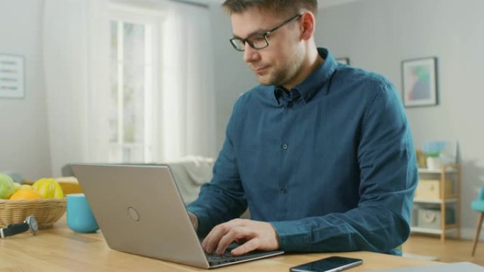 穿着Galsses和海军蓝衬衫的男人坐在家里的桌子旁，在他的现代银色笔记本电脑上工作。智能手机躺在他