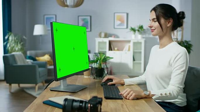 美丽的年轻女摄影师坐在舒适公寓的办公桌前，在绿色模拟屏幕个人电脑上工作。