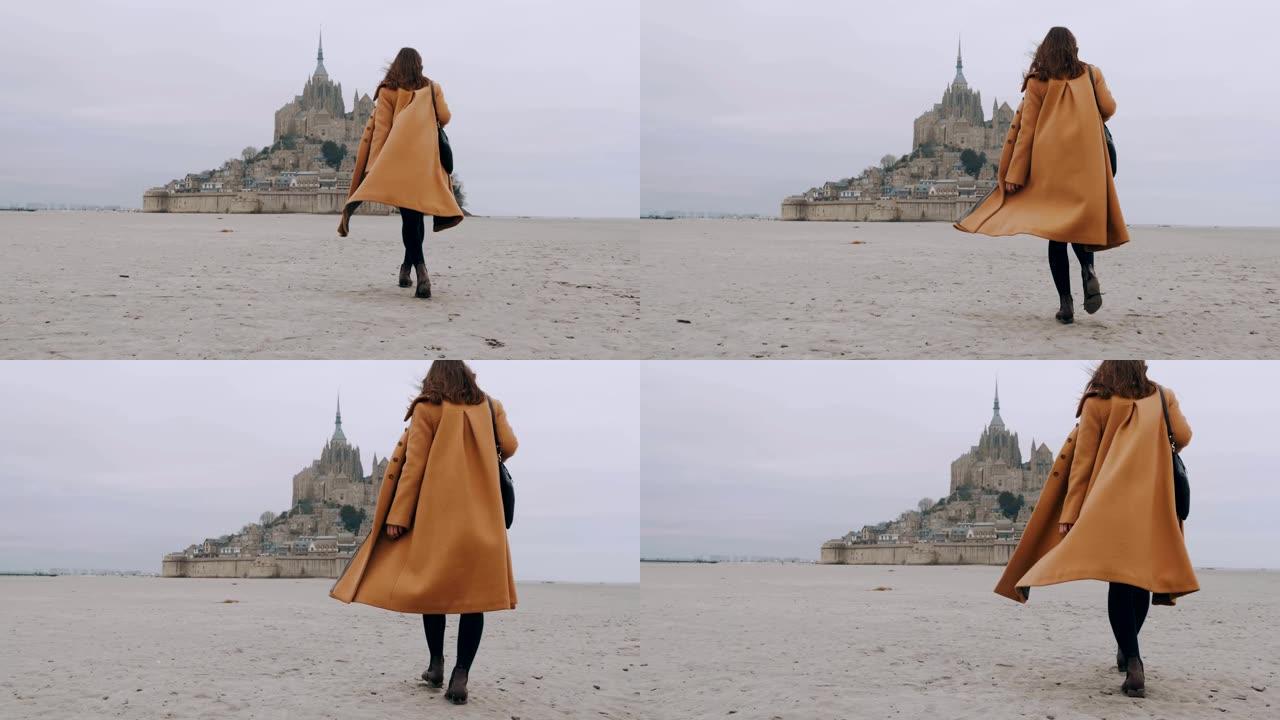 后视图放大时尚旅游女性在史诗圣米歇尔山城堡慢动作中缓慢行走在海沙上。