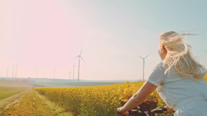 超级SLO MO - TIME WARP EFFECT女人骑着自行车沿着油菜籽，远处有风力涡轮机