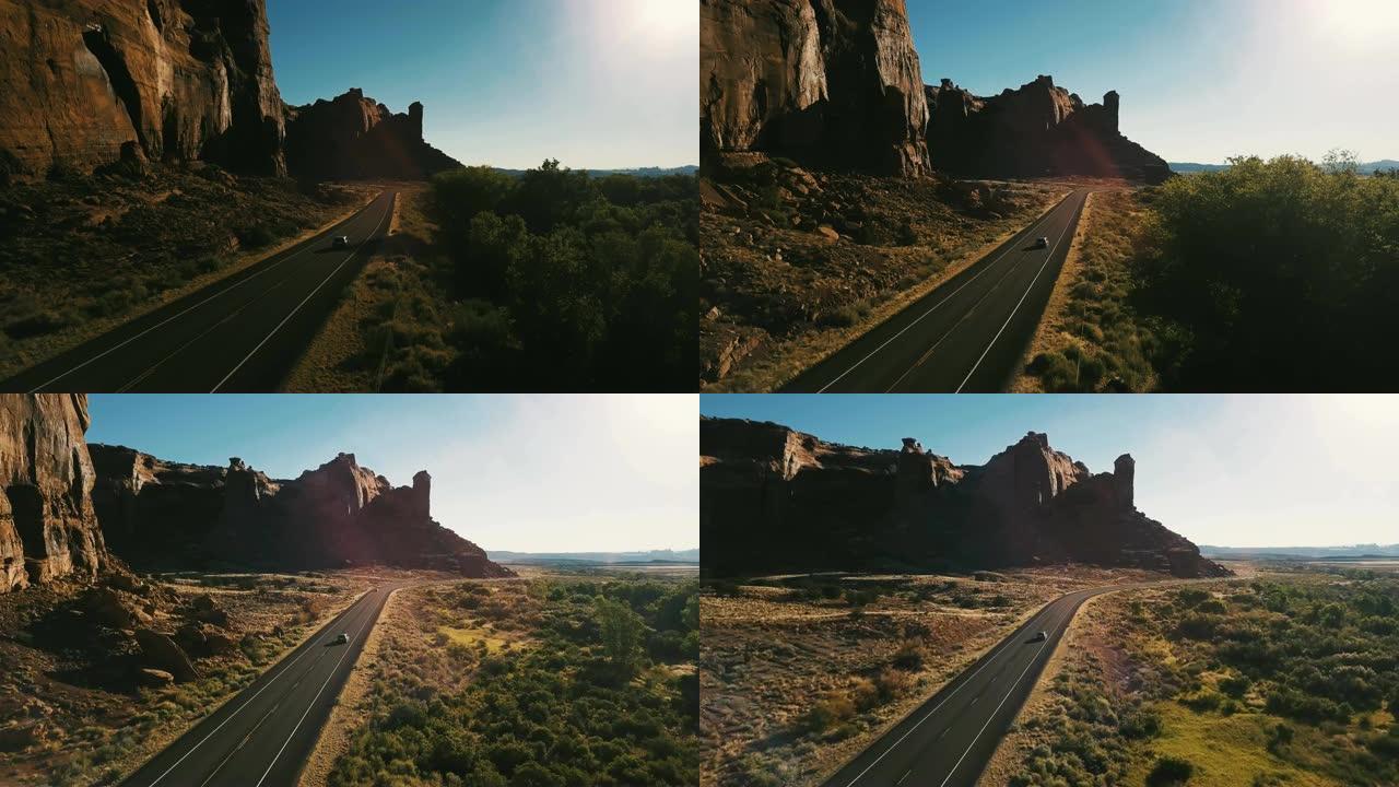 无人机摄像机跟随汽车在美国日落时沿着小沙漠公路行驶，靠近令人惊叹的陡峭峡谷山脊。