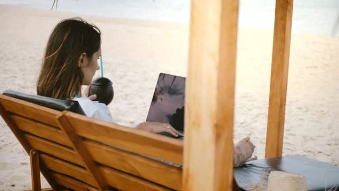 在海滩上的躺椅上使用笔记本电脑喝酒的聚焦成功工作狂女商人的惊人背景照片。