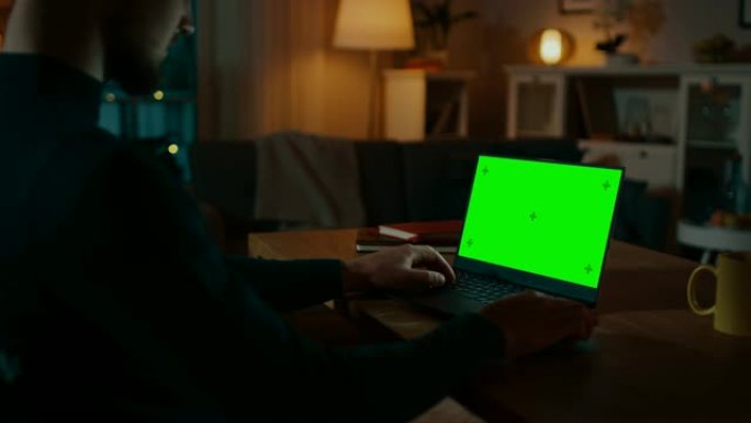 肩膀上的镜头: 坐在办公桌前的男人在带有绿色色度键屏幕的笔记本电脑上工作。深夜在她的客厅里，男人使用