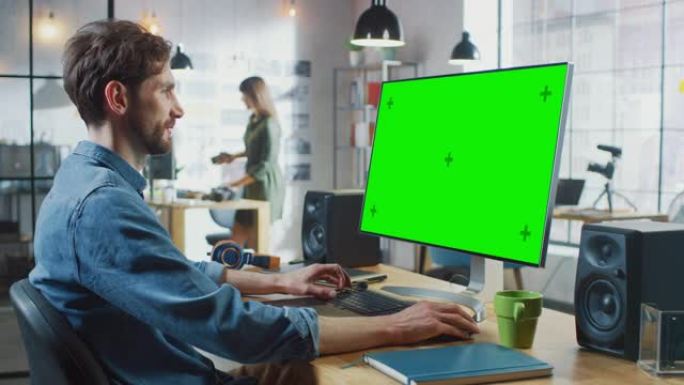 留着胡须和牛仔裤衬衫的男性视频编辑器在他的个人计算机上工作，并带有绿色大屏幕模拟显示。他在一个很酷的