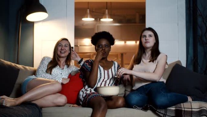 快乐的年轻多民族女性朋友坐在一起在家看电影，一起微笑着吃零食慢动作。