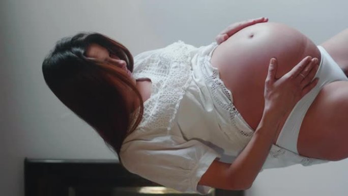 一个穿着白色连衣裙的年轻孕妇的真实垂直镜头正在抚摸她的肚子，她早上刚在卧室醒来